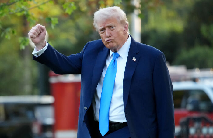 Donald Trump in a dark suit and blue tie raising his fist outdoors, related to paying Americans 2000 from tariff revenue.