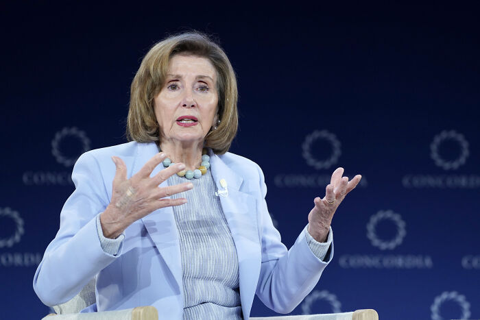 Nancy Pelosi speaking at an event, wearing a light blue blazer and gesturing with her hands against a dark blue background.