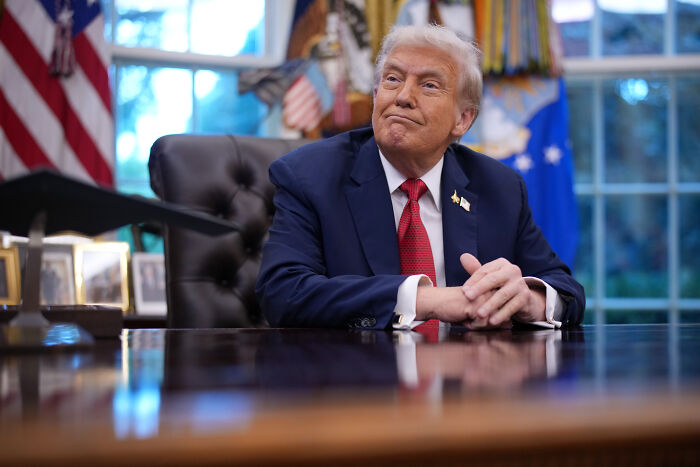 Donald Trump sitting at the desk in the Oval Office during European summit, smiling amid geographic gaffe mockery.