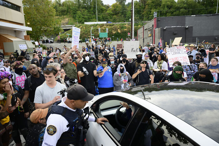 Portland protester surrounded by crowd and police, some holding signs during a viral protest scene in the city.