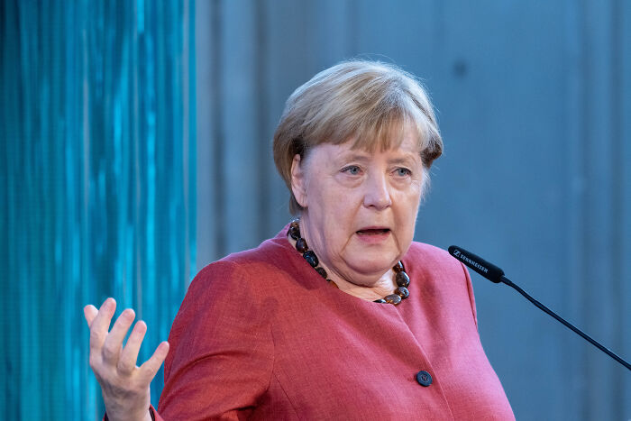 Angela Merkel speaking at a podium, discussing Poland and Baltic states fueling Putin&rsquo;s war in Ukraine.