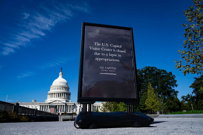 Sign outside US Capitol Visitor Center about closure, with Capitol dome and clear blue sky in background, related to Trump and Russ Vought.