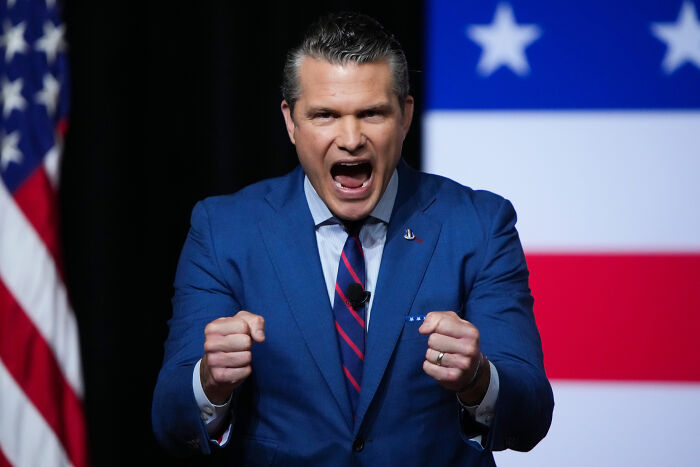 Man in a blue suit speaking passionately with clenched fists in front of a blurred American flag backdrop.