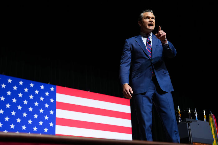 Man in a blue suit speaking on stage with a large American flag backdrop during a military training grounds discussion.