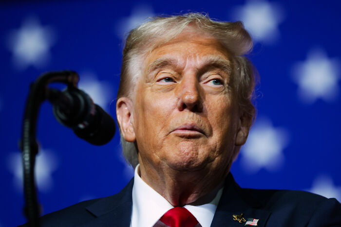 Donald Trump speaking at a podium, with a blurred American flag background, related to House Speaker Mike Johnson.