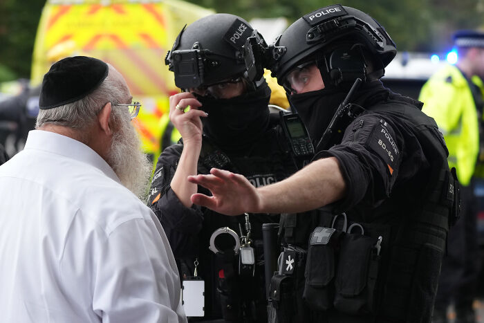 Police officers in tactical gear speaking with a man outside Manchester synagogue after terror attack on Yom Kippur.