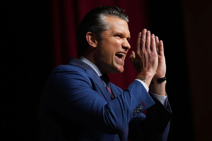 Pete Hegseth speaking passionately at an event, wearing a blue suit against a dark red background.