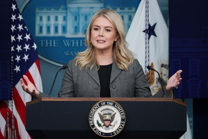 Karoline Leavitt speaking at a podium with US flags behind her, defending Trump&rsquo;s AI medbed video.