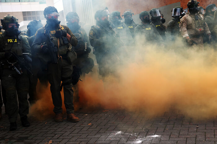 Portland protester refusing to walk being rolled away on a cart amid police and orange smoke during a protest.