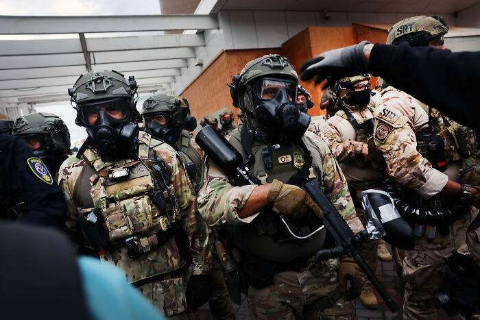 Law enforcement in tactical gear and gas masks during a tense standoff linked to MAGA and anti-ICE sentiments.
