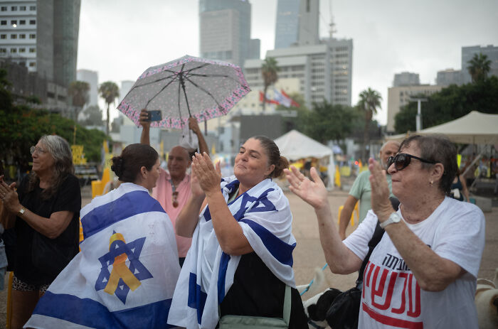 People wrapped in Israeli flags gathering outdoors, expressing hope and doubt about Hamas hostage release and Israel ceasefire commitment.