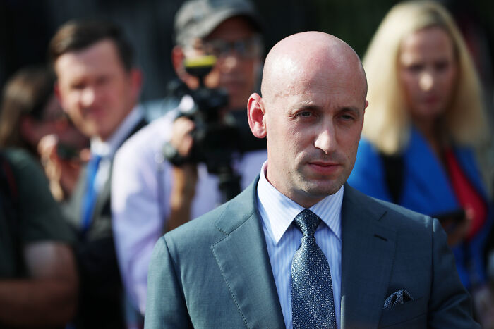 Bald man in a suit and tie at a public event, with blurred people and a camera in the background.