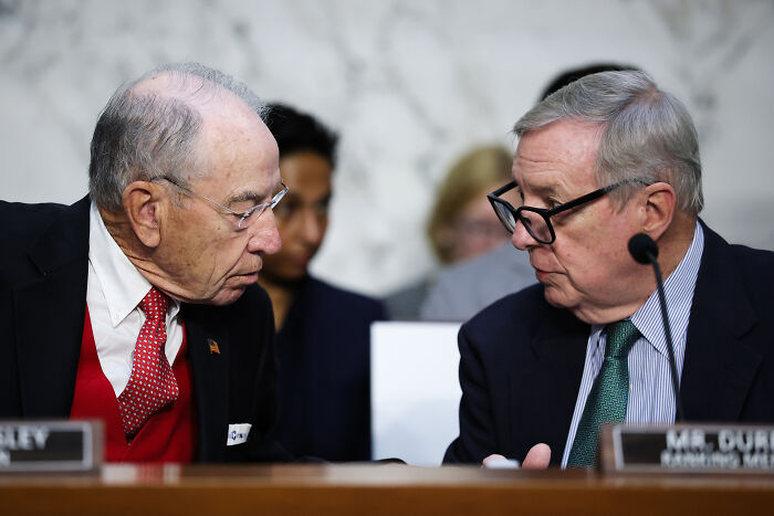 Two older men in suits and glasses engaged in discussion during a formal hearing on Trump photos linked to Epstein investigation
