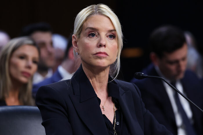 Bondi wearing a black suit, sitting at a hearing, dodging questions about alleged Trump photos linked to Epstein investigation.