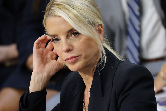 Pam Bondi in a black blazer, sitting thoughtfully during a formal event amid a serious atmosphere.