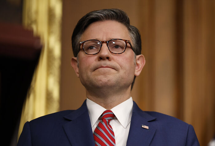 Mike Johnson speaking at a podium wearing glasses and a suit, discussing role model choice for Super Bowl replacement.