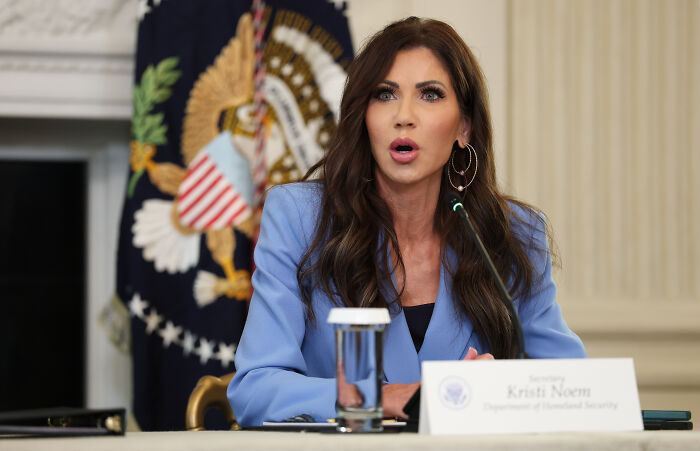 Kristi Noem speaking at a government event with a blue blazer and earrings, U.S. flag in the background.