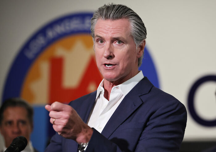 Gavin Newsom speaking at a podium during a press event, related to trolling Trump with a Time Magazine cover edit.
