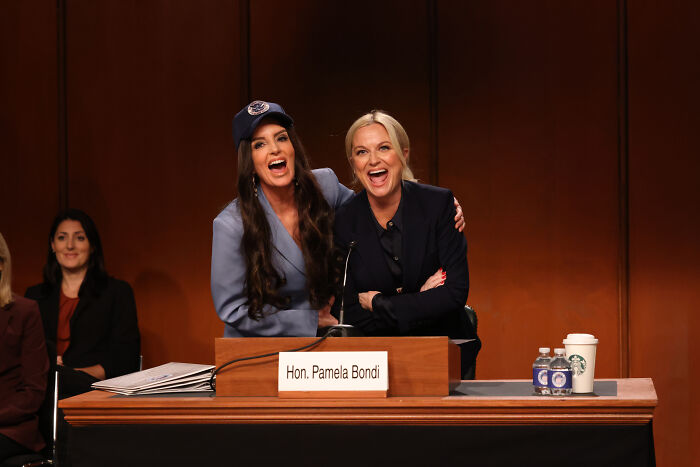 Two women smiling and posing at a panel with a sign reading Hon. Pamela Bondi after Homeland Security anti-ICE comments.