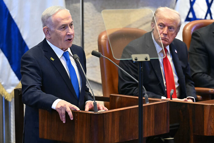 Benjamin Netanyahu speaking at the Knesset podium with Donald Trump seated nearby during a formal event.
