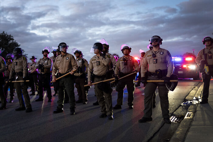 Homeland Security officers in riot gear standing in a line on a street with flashing police lights at dusk.