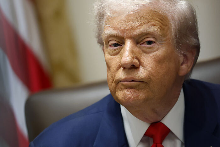 Close-up of a man in a blue suit and red tie with an American flag blurred in the background, related to White House topics.