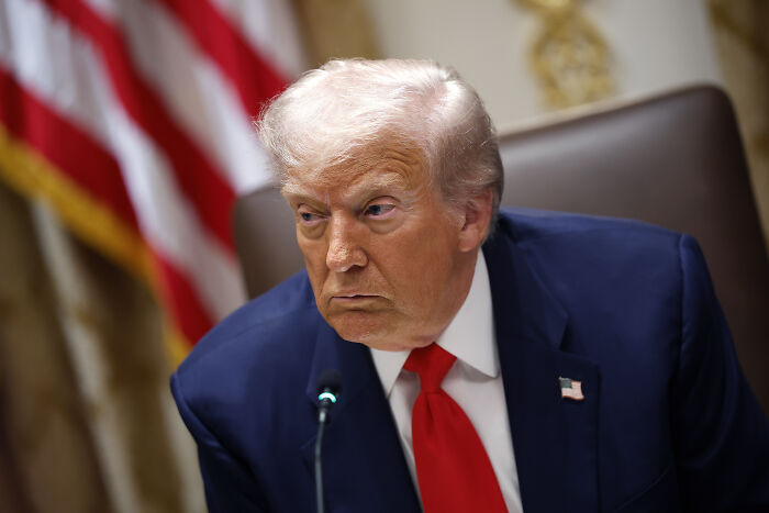 Former president in a dark suit speaking at a meeting, with an American flag blurred in the background.