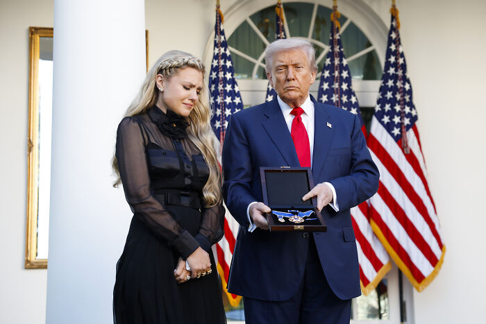 Man in suit holding medal next to woman in black dress with multiple American flags in the background at formal event.