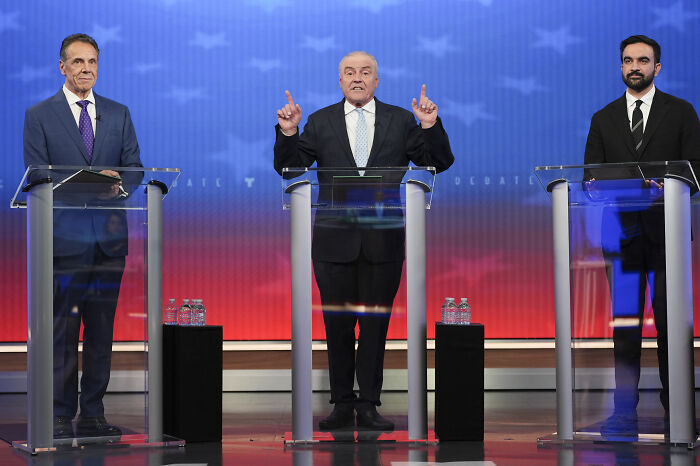 Three NYC mayoral candidates including Zohran Mamdani speaking at a debate stage with clear podiums and a red backdrop.