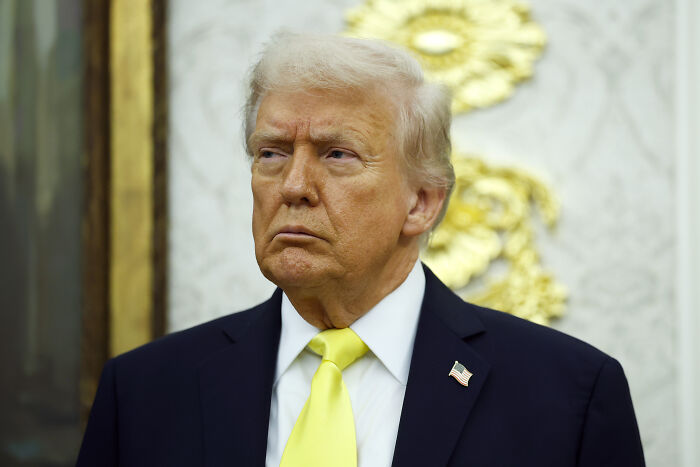 Donald Trump in a suit with a yellow tie, standing indoors with gold and white decorative background, focused expression.