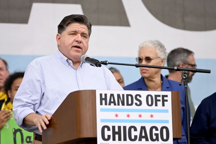 Man speaking at a podium with a hands off Chicago sign, addressing a crowd during a political event related to John Cusack.
