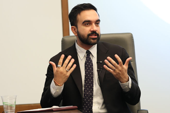 Zohran Mamdani speaking at a panel, wearing a suit and tie, leading NYC mayoral race in recent poll results.