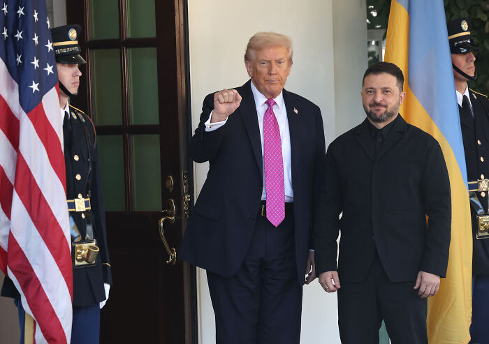 Donald Trump with clenched fist standing next to Volodymyr Zelenskyy outside with flags and guards present.