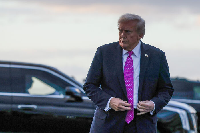 Former president in a dark suit and pink tie standing outside near black vehicles referencing authoritarian takeover warnings.