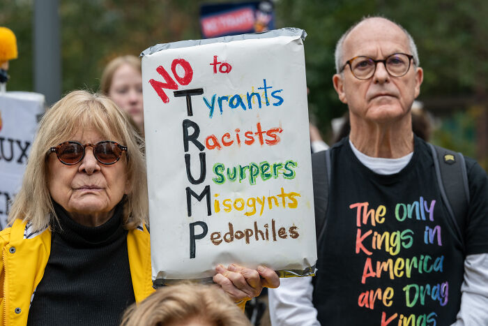 Two protesters holding a sign opposing Trump with colorful messages on racism and misogyny during a public demonstration.