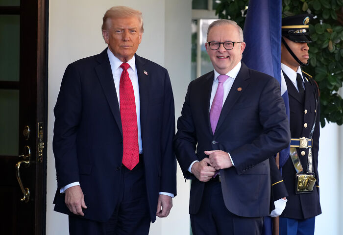 Donald Trump standing with a visitor in a suit and red tie outside the White House with a security guard nearby.