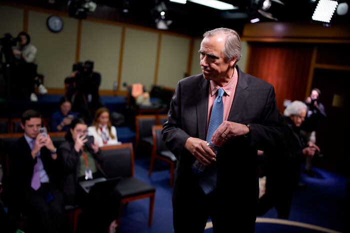 Senator speaking in a formal setting, holding a water bottle, delivering a floor speech warning of authoritarian takeover.