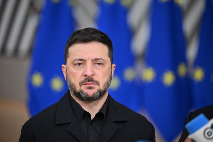 Volodymyr Zelenskyy standing solemnly with European Union flags in the background during a public appearance.