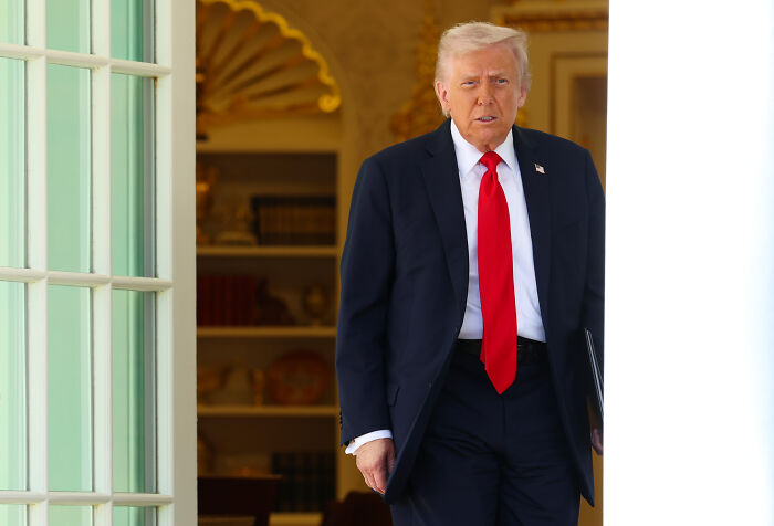 Donald Trump in a dark suit and red tie walking out of a doorway, related to $230 million compensation bid against DOJ.