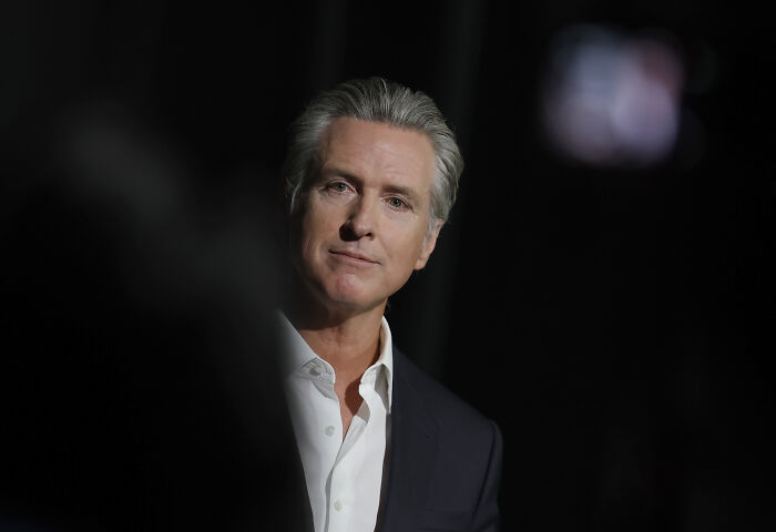 Gavin Newsom wearing a dark suit and white shirt, speaking at an event with a serious expression on a dark background