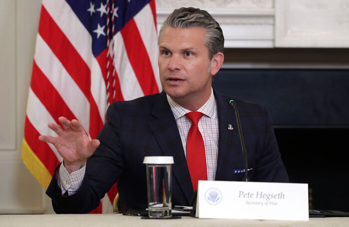 Pete Hegseth speaking at a briefing, wearing a dark suit and red tie, with an American flag in the background.
