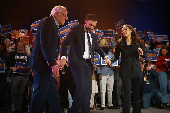 AOC at a campaign event holding hands with two men, with supporters holding signs in the background.