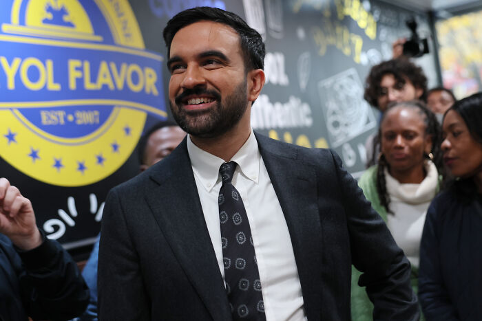 Zohran Mamdani, NYC mayoral frontrunner, smiling and wearing a suit at an indoor event with people in the background.