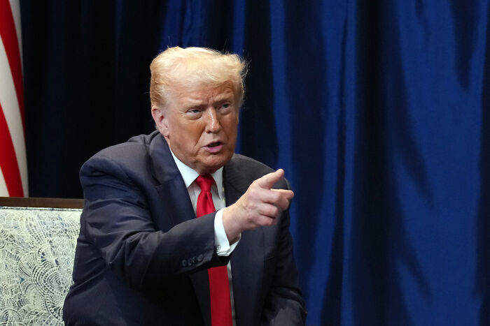 Former President Donald Trump in a dark suit and red tie pointing during a conversation against a blue curtain background related to Stephen King World Series post.
