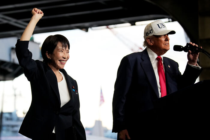 Donald Trump speaking at a podium aboard an aircraft carrier, with a woman raising her fist beside him.
