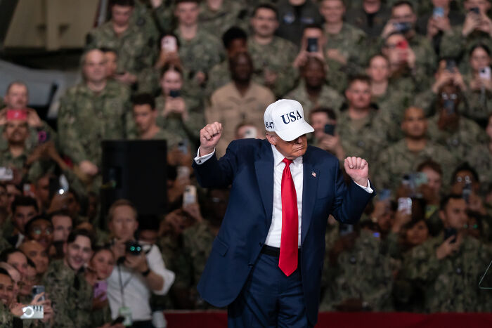 Donald Trump wearing a USA hat and red tie, gesturing during a speech aboard an aircraft carrier with military personnel watching.