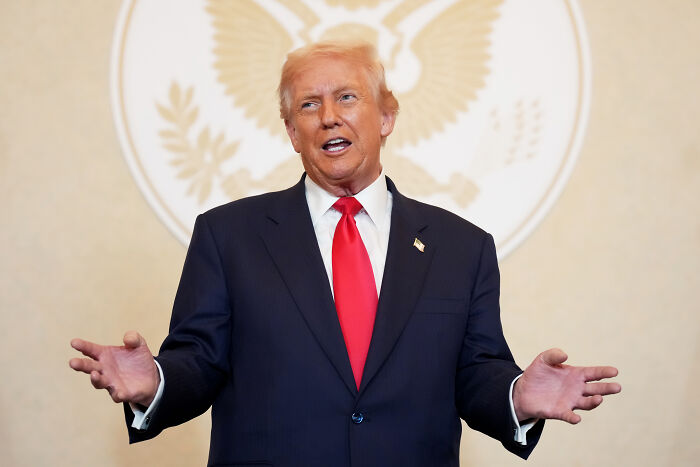 Donald Trump in a dark suit with a red tie speaking and gesturing during an Asia summit event with a presidential emblem backdrop.