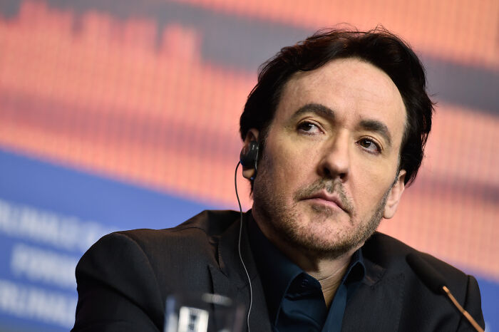 John Cusack wearing a black suit and earpiece, speaking at a formal event with an orange and blue background.