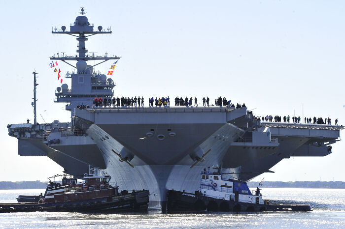 U.S. Navy&rsquo;s largest aircraft carrier sailing with crew on deck, escorted by tugboats in open water near the Caribbean.