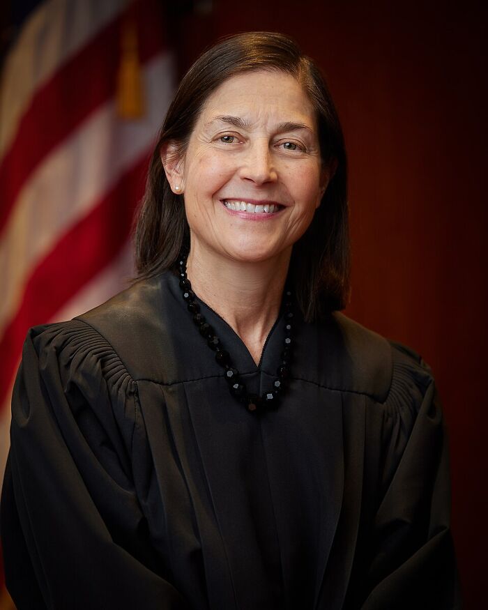Smiling woman in judicial robe posing in front of a blurred American flag in an official portrait setting.
